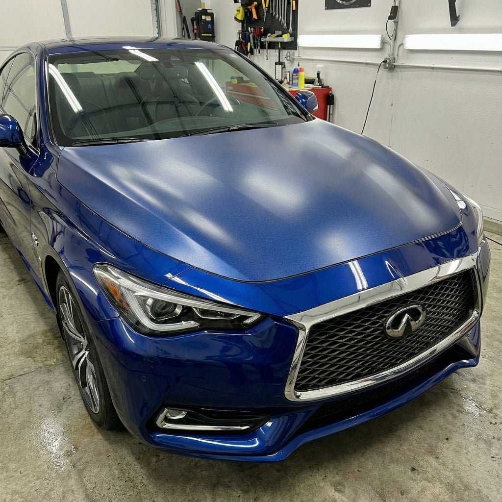 A shiny blue Infiniti sedan sits in a garage, with detailing supplies and Transparent Matte Sparkle SelfRepair Nano-Coated PPF displayed on the wall and counter in the background.