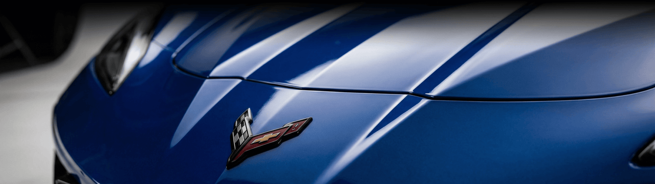 Close-up of the front hood of a blue Chevrolet Corvette, featuring the distinctive Corvette emblem and sharp body lines reflecting light.