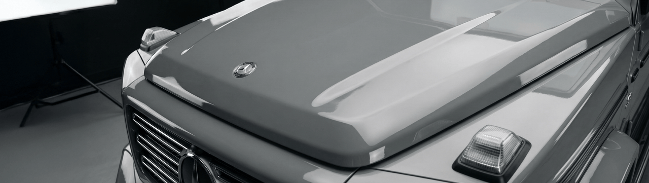 Close-up view of the front hood and headlights of a gray Mercedes-Benz G-Class SUV in a studio setting, with part of a lighting setup visible on the left.