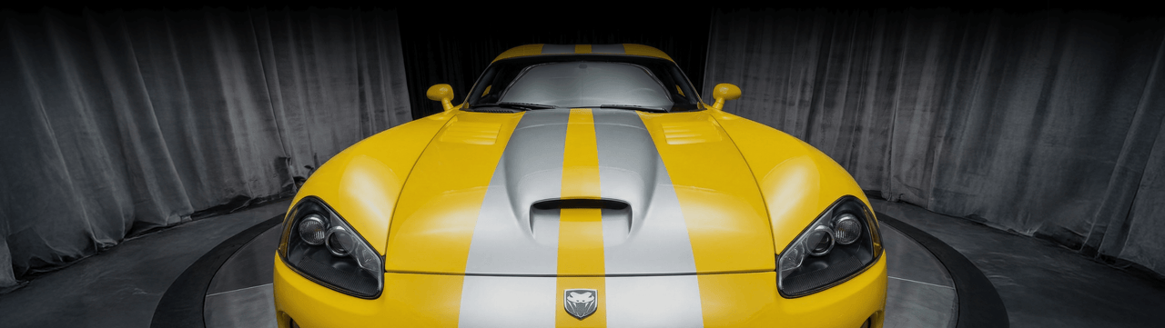 A yellow sports car with silver racing stripes is parked indoors in front of gray curtains, shown from a low, head-on angle. The car features aggressive headlights and a central hood scoop.