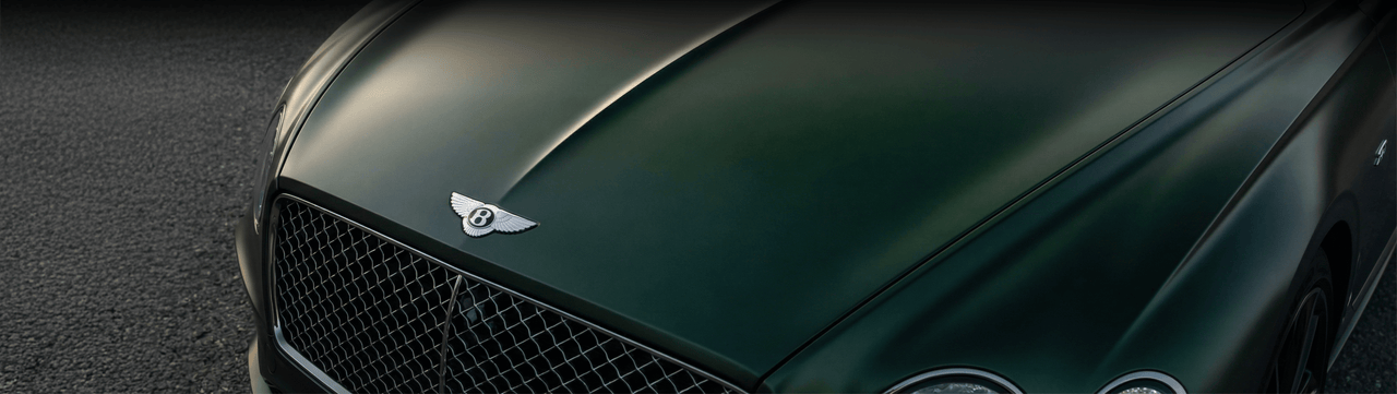 Close-up view of the front hood and grille of a dark green Bentley car, featuring the Bentley emblem and part of the vehicle’s headlights, with an asphalt surface in the background.