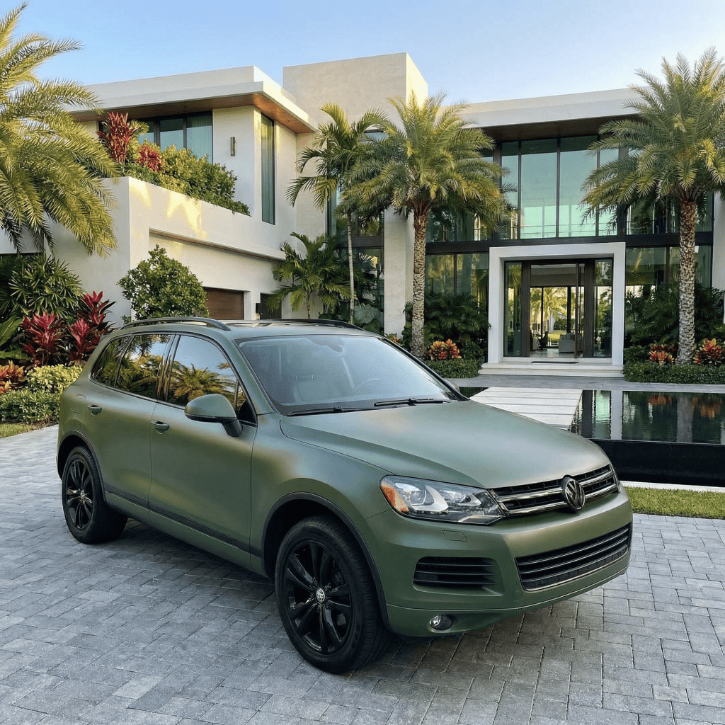 A Volkswagen SUV wrapped in Satin Army Green SelfRepair Nano-Coated PPF is parked on a stone driveway before a modern, luxurious house with palm trees and lush landscaping.