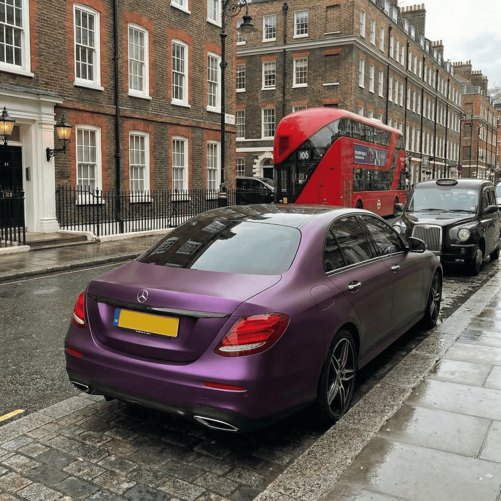 A Mercedes-Benz sedan wrapped in Satin Black Pinot Purple SelfRepair Nano-Coated PPF is parked on a wet city street near a black taxi and red double-decker bus. Brick buildings with white trim stand in the cloudy background.
