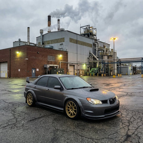 A Subaru sedan in Satin Gray Meteorite Matte with gold wheels is parked on wet pavement by a factory under clouds, protected by Satin Gray Meteorite SelfRepair Nano-Coated PPF.