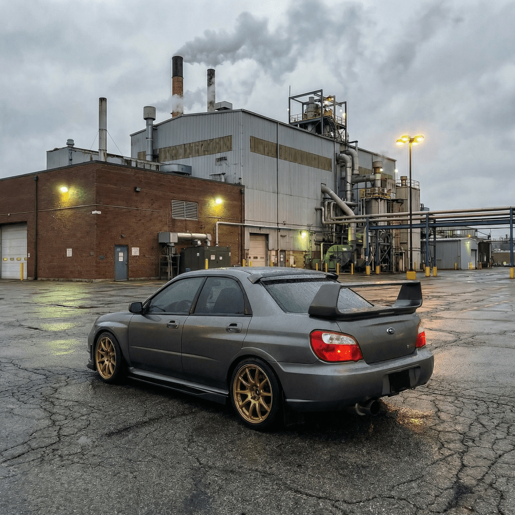A Satin Gray Meteorite Matte sports sedan with gold wheels and a large rear spoiler is parked on cracked pavement before a factory, its finish protected by Satin Gray Meteorite SelfRepair Nano-Coated PPF under cloudy skies.