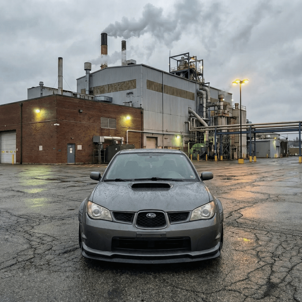 A Subaru sedan protected by Satin Gray Meteorite SelfRepair Nano-Coated PPF is parked outside an industrial factory on a cloudy day, with smoke from chimneys and yellow lights reflecting off the wet, cracked pavement.