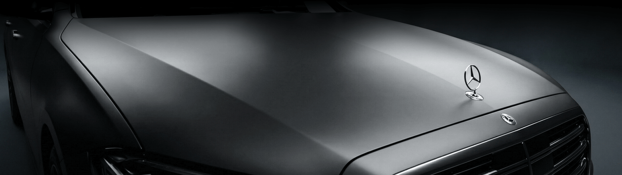 Close-up of the front hood of a sleek, matte-finished black Mercedes-Benz car, featuring the iconic three-pointed star emblem and part of the front grille, photographed against a dark background.