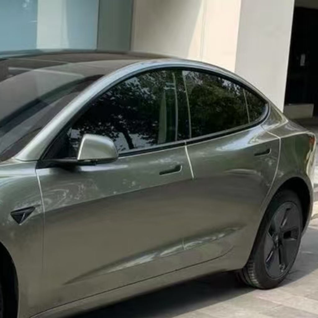 An Alpine Green Tesla Model 3 with Alpine Green Liquid Metallic Glossy SelfRepair Nano-Coated PPF is parked by a modern building. Its tinted windows and advanced nano-coating reflect the nearby trees.