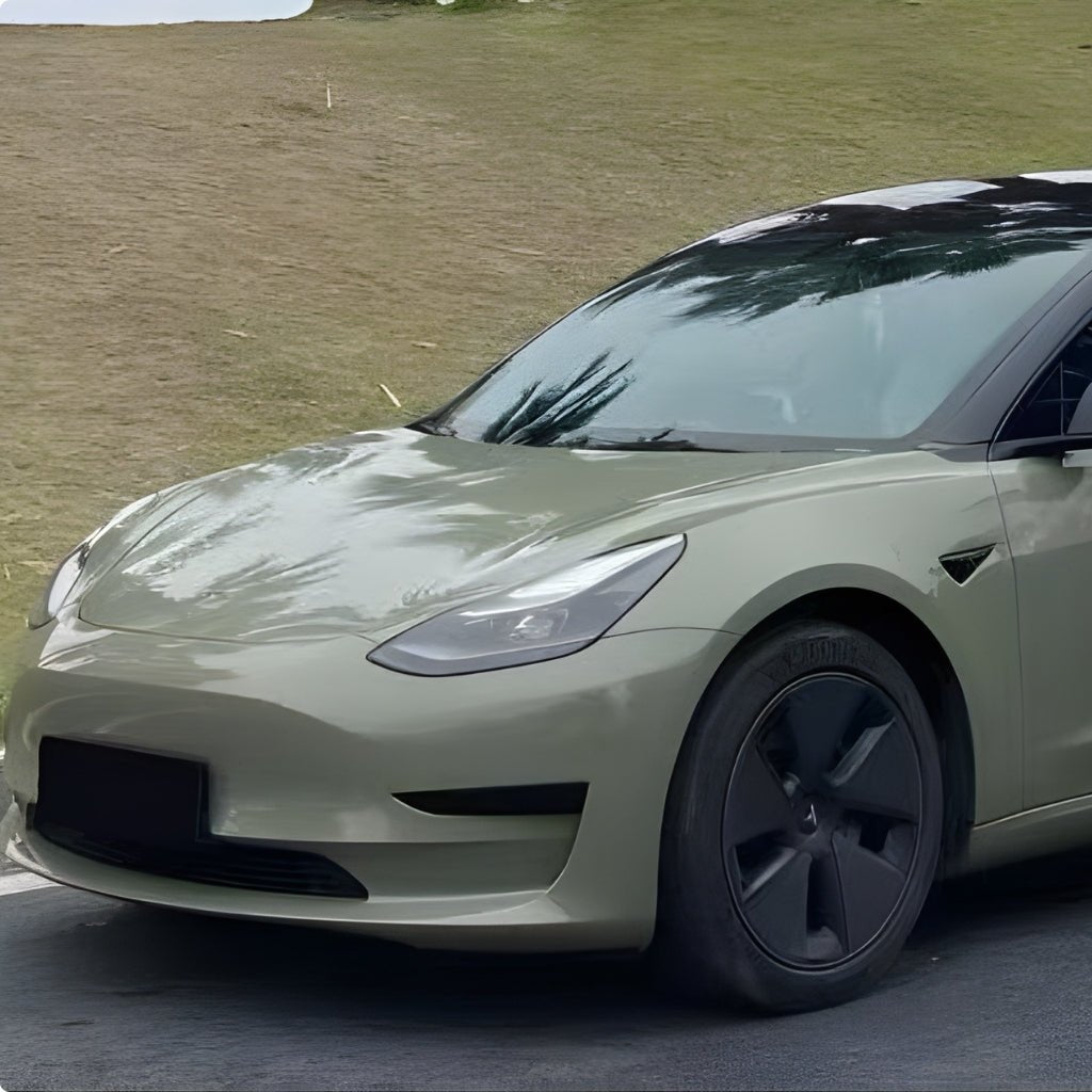 A Tesla Model 3 in light army green with black wheels, protected by Army Green Glossy SelfRepair Nano-Coated Nexthor™ PPF, is parked on a road before a grassy hillside.