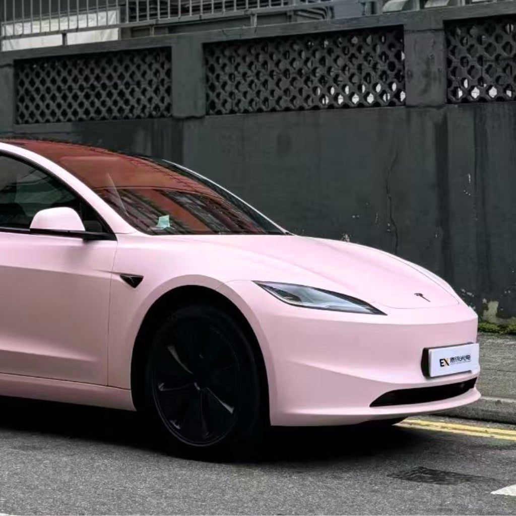 A light pink Tesla Model 3 with black wheels, protected by Baby Pink SelfRepair Nano-Coated PPF for extra scratch protection, is parked near a building with a dark fence and concrete wall.
