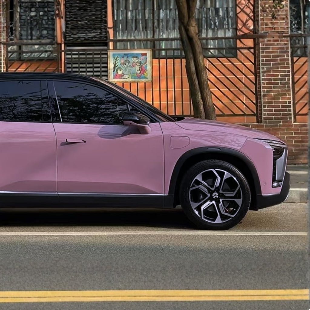 A pink SUV wrapped in Blush Mauve Glossy SelfRepair Nano-Coated PPF is parked by a brick building with metal fencing, a large window, and wall art behind the fence. A tree stands nearby.