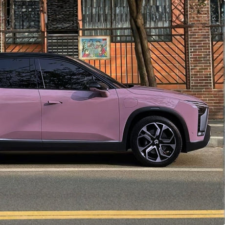 A pink SUV wrapped in Blush Mauve Glossy SelfRepair Nano-Coated PPF is parked by a brick building with metal fencing, a large window, and wall art behind the fence. A tree stands nearby.