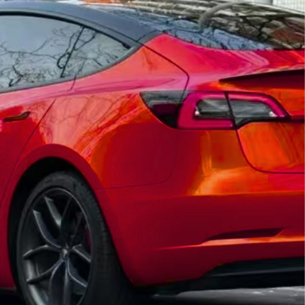 A rear-side view of a shiny red car highlights its back wheel, tail light, and trunk, shielded by Burnt Orange Glossy SelfRepair Nano-Coated PPF. Reflections of trees and a building shimmer on its sleek surface.