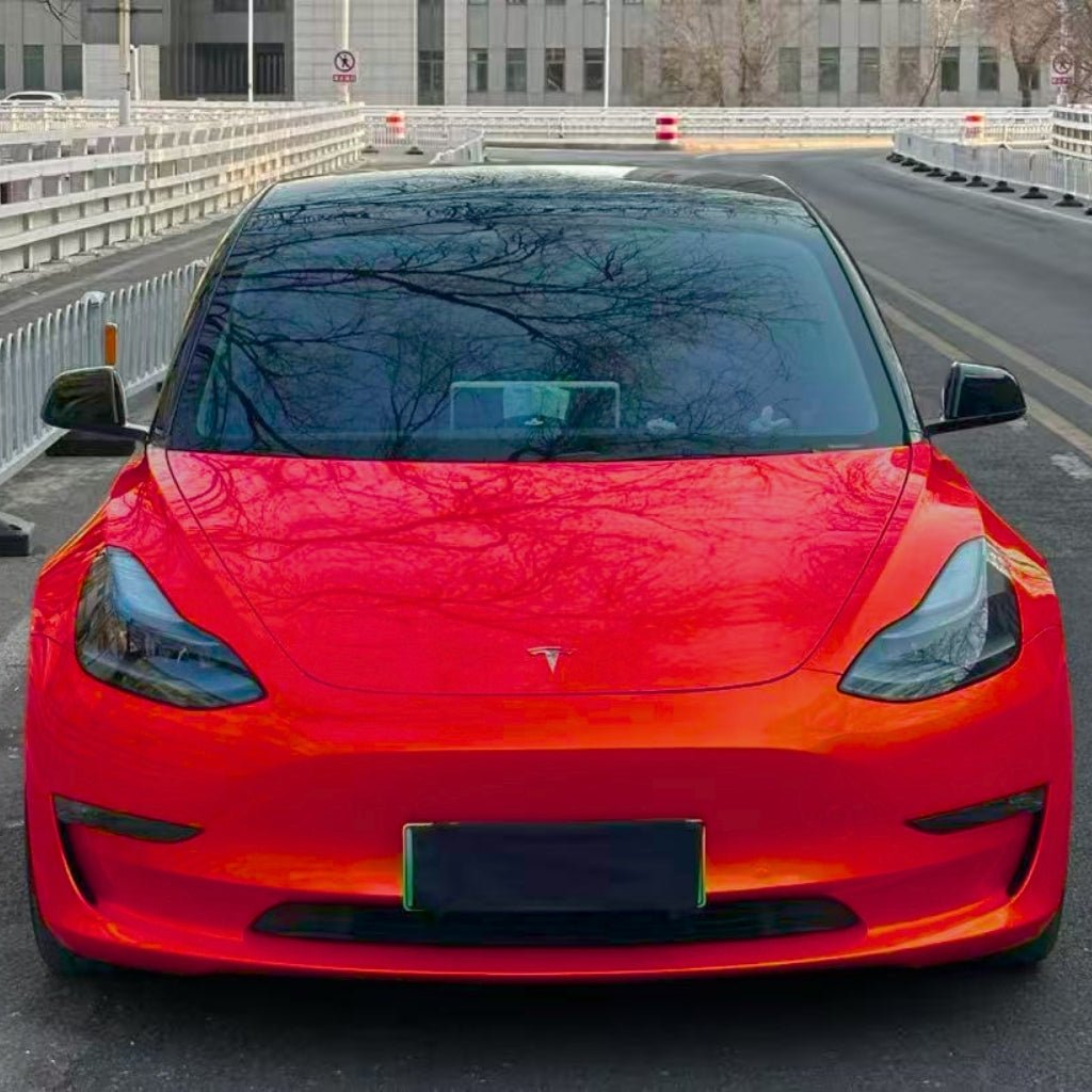 A bright red Tesla Model 3 with Burnt Orange Glossy SelfRepair Nano-Coated PPF is parked on a city street, facing forward. Leafless tree branches reflect on its windshield, and white barriers line both sides of the road.