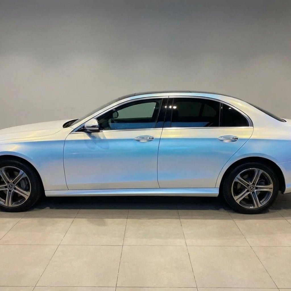 A silver luxury sedan, wrapped in White Diamond Blue Glossy SelfRepair Nano-Coated PPF, is parked indoors on a tiled floor, positioned sideways against a plain light gray wall.