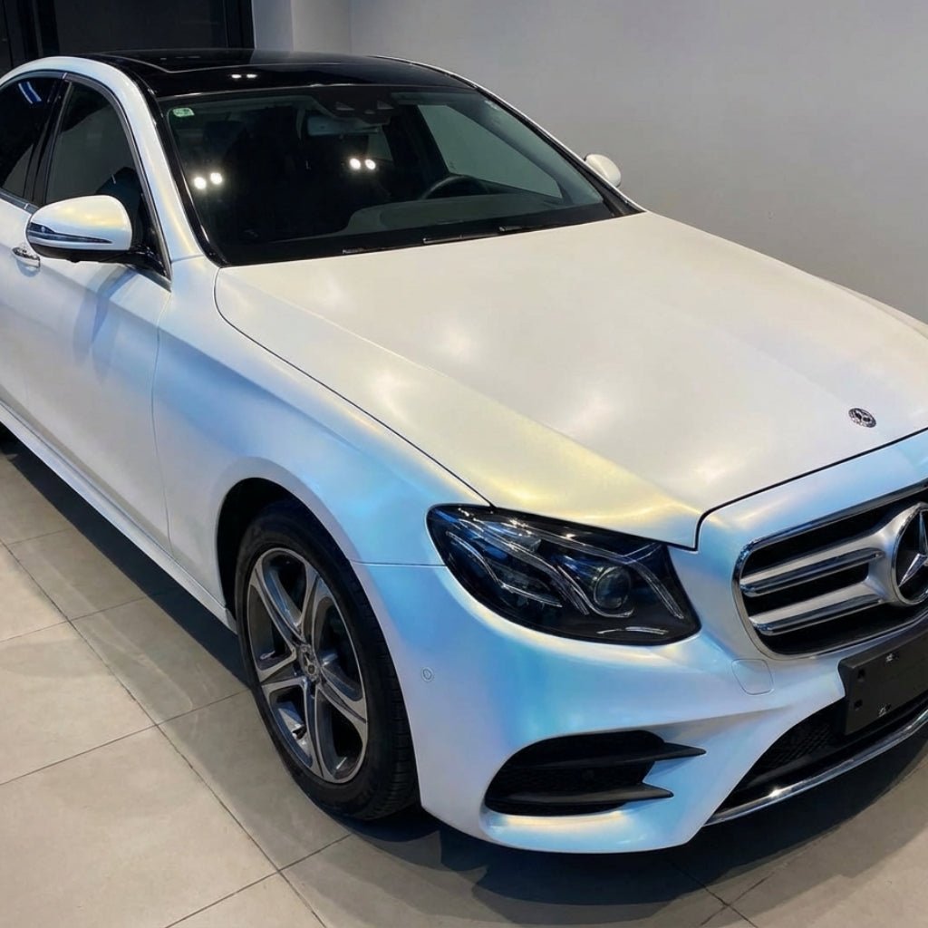 A white Mercedes-Benz sedan is parked indoors on a tiled floor, its pristine look enhanced by White Diamond Blue Glossy SelfRepair Nano-Coated PPF for superior protection and shine. The headlights are off, and the car looks clean and well-maintained.