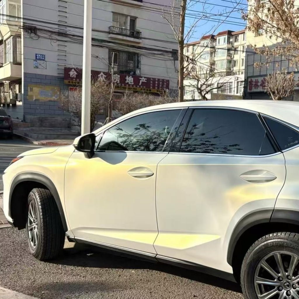 A white SUV with White Diamond Gold Glossy SelfRepair Nano-Coated PPF is parked by apartment buildings on a sunny day, with tree branches and buildings reflected in its tinted windows.