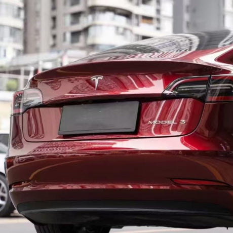 A close-up of the rear of a Tesla Model 3 wrapped in Dragon Blood Red Liquid Metallic Glossy SelfRepair Nano-Coated PPF, parked outdoors with modern apartment buildings blurred behind. The car’s badge and logo appear above the license plate.