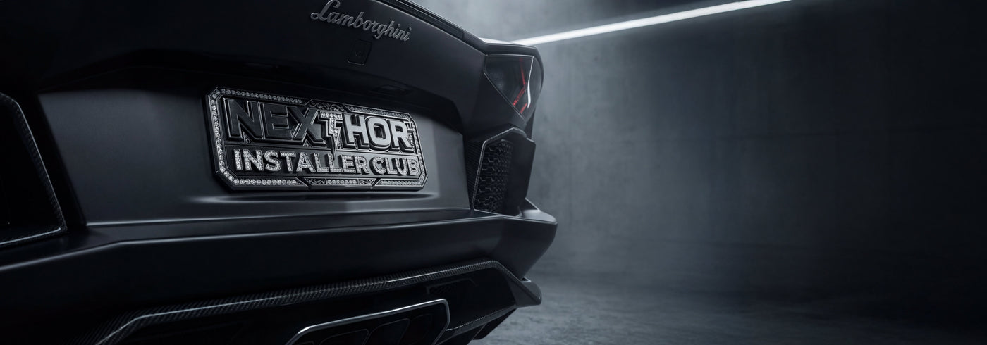 Close-up of the rear of a matte black Lamborghini in a dimly lit garage, featuring a custom license plate that reads NEXTLEVEL INSTALLER CLUB.