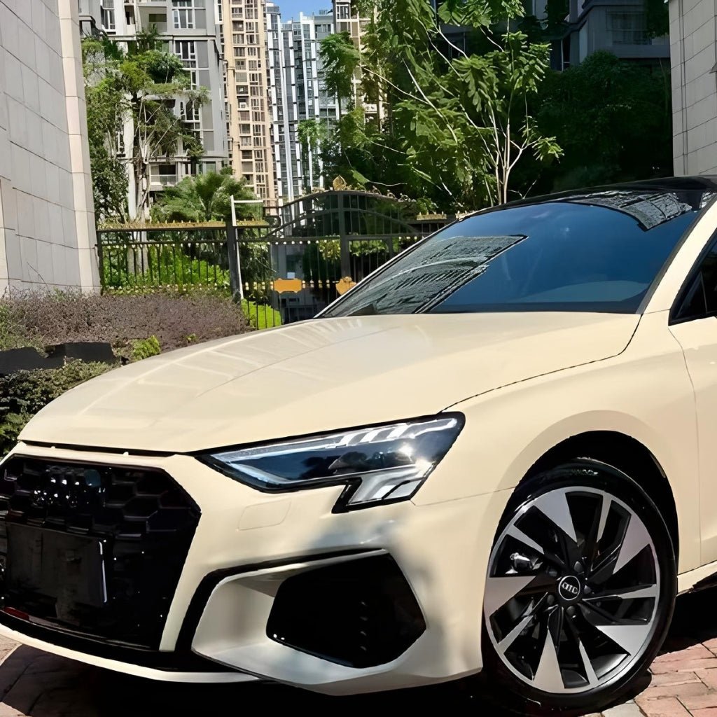 A cream Audi sedan with Khaki Milan Glossy SelfRepair Nano-Coated PPF for maximum body protection is parked on a brick driveway in front of modern apartments and green trees.