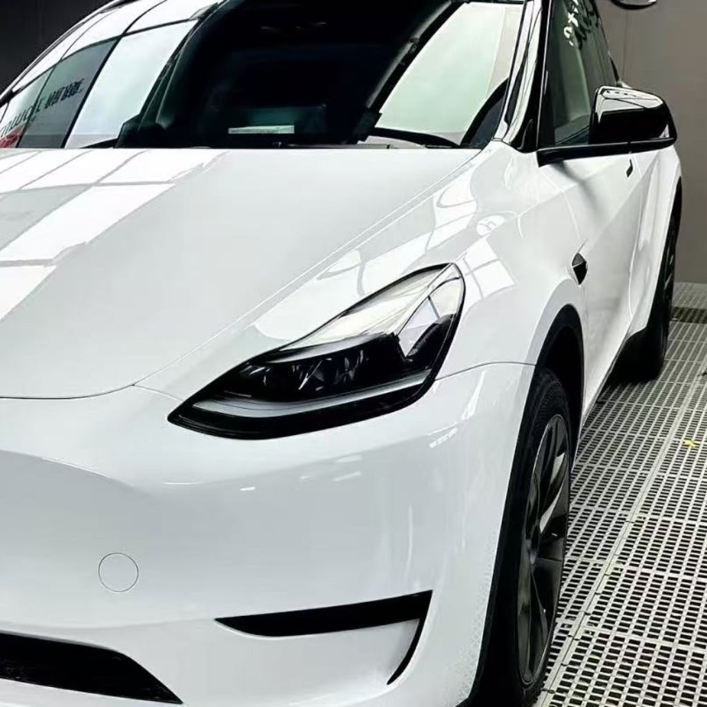 A close-up of the front right side of a white Tesla Model Y highlights its sleek headlights, modern curves, and flawless Metallic White Glossy SelfRepair Nano-Coated PPF, all showcased inside a well-lit garage.