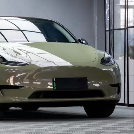 A partial view of a vojenská zelená Tesla Model 3 with a blank license plate, parked indoors on a patterned floor, its shiny surface reflecting windows and protected by Military Green Glossy SelfRepair Nano-Coated PPF.
