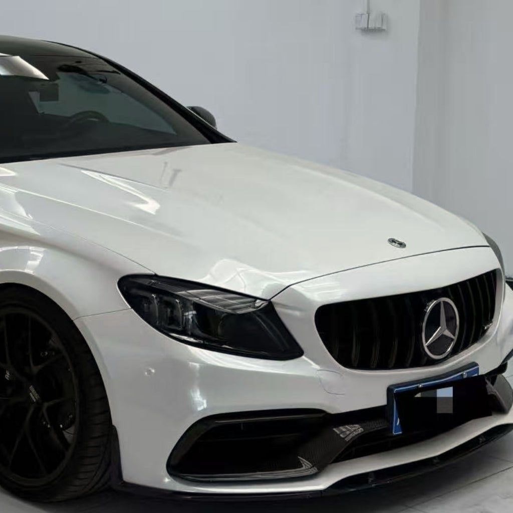 A white Mercedes-Benz parked indoors showcases the Arctic White Sparkle Glossy SelfRepair Nano-Coated PPF for ochrana karosérie. The front grille, Mercedes emblem, headlights, and part of a black wheel stand out against a clean white background.