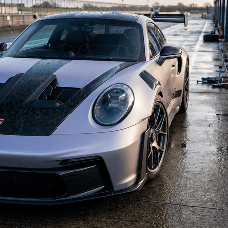 A high-performance sports car with a matte pink and black, carbon-inspired finish features Forged Carbon Graphite Self-Healing Nano-Coated PPF, aerodynamic vents, racing wheels, and a large rear wing on a wet racetrack.