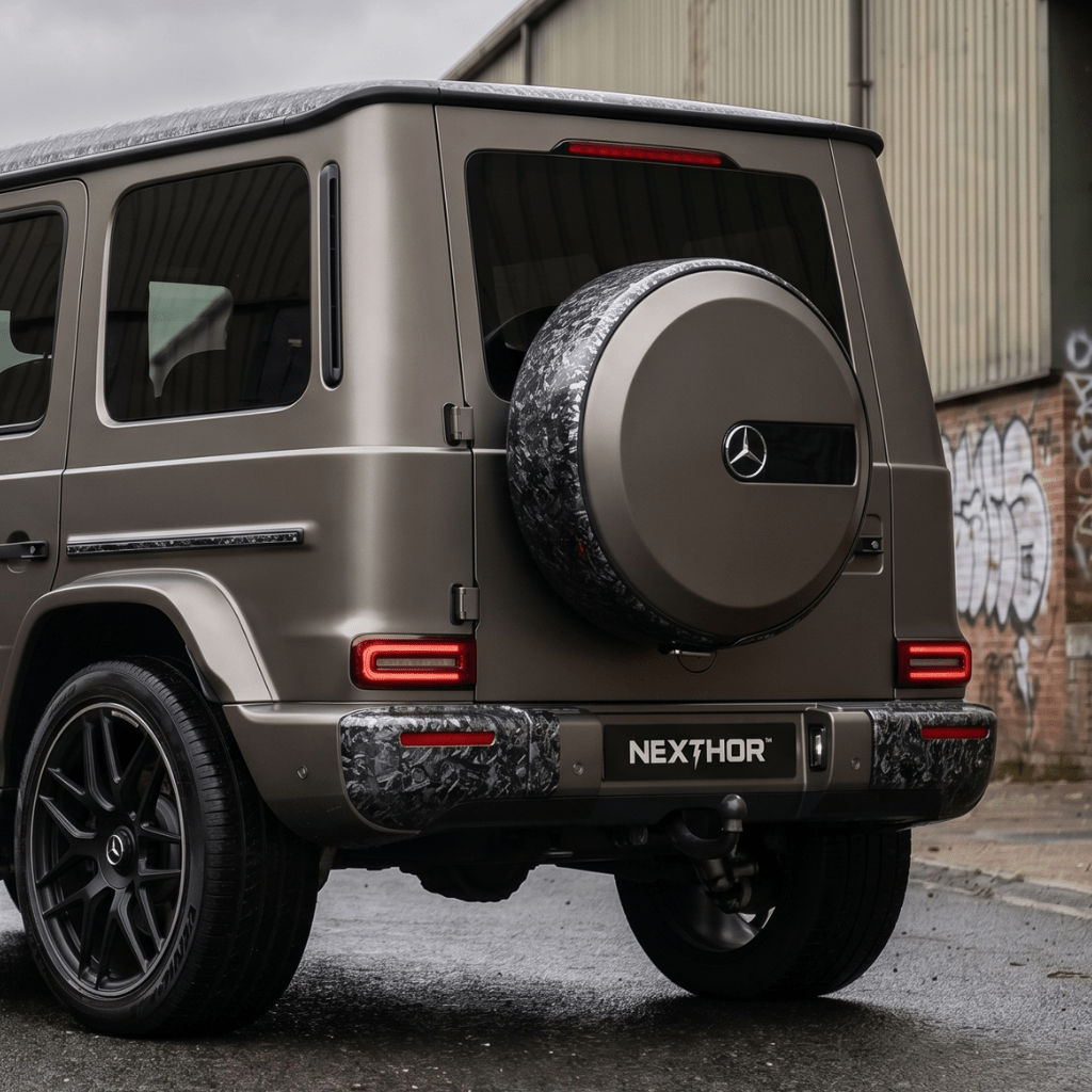 A matte gray Mercedes-Benz SUV is parked on a wet street near an industrial building, featuring a bold Forged Carbon Graphite Self-Healing Nano-Coated PPF finish and a NEXTHOR plate for a striking rear view.