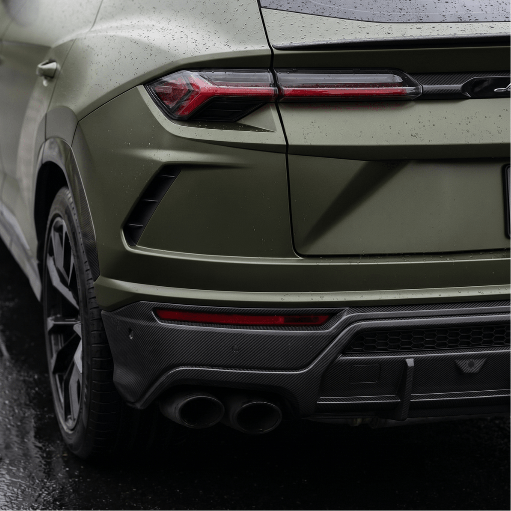Close-up of a matte green luxury SUV with Matte Carbon Fiber SelfRepair Nano-Coated PPF, showing raindrops on the surface, modern tail lights, dual exhausts, and black trim glistening on wet pavement.