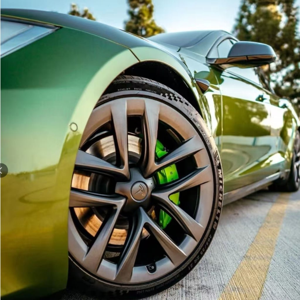 Close-up of a green Tesla’s front wheel with black and silver rims and green brake calipers—protected by Blue Gold Green Chameleon SelfRepair Nano-Coated PPF—parked on a concrete road with yellow lines, blurred trees in the background.