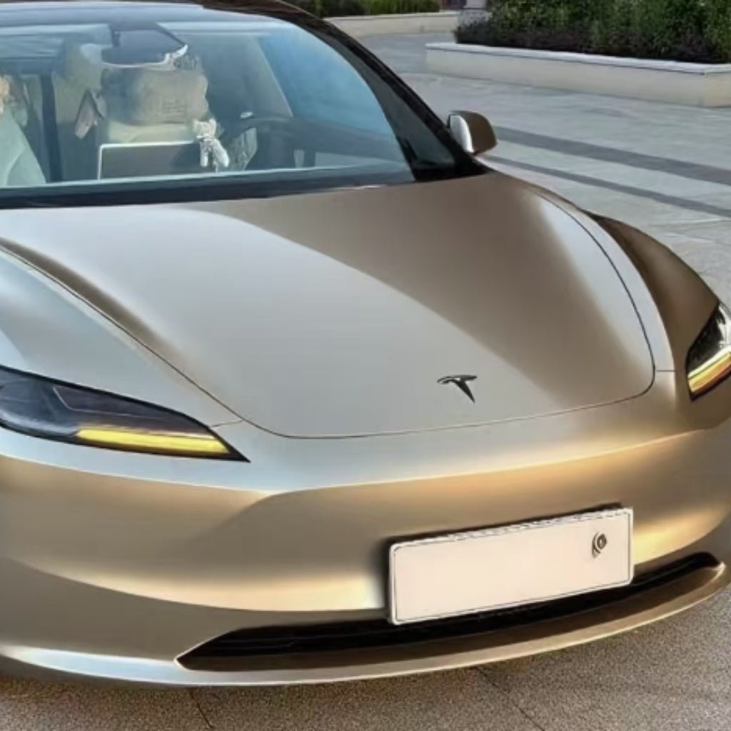 A sleek Tesla featuring Champagne Gold Liquid Matte Metallic SelfRepair Nano-Coated PPF is parked outdoors on pavement, with modern landscaping and greenery in the background. The car’s front license plate is blank.
