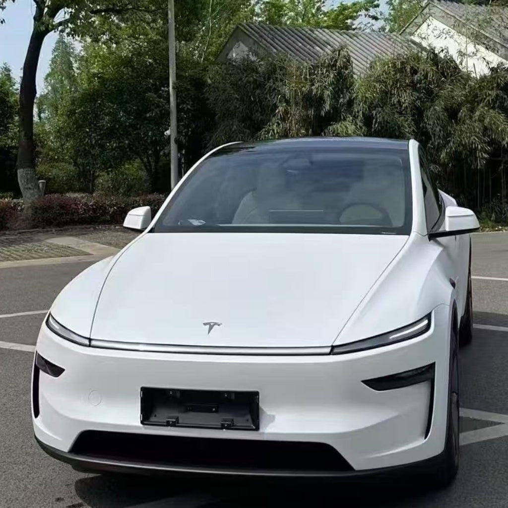 A sleek white Tesla with White Diamond Glossy SelfRepair Nano-Coated PPF is parked on a street near greenery and modern buildings, viewed from the front at an angle under a clear sky surrounded by trees.