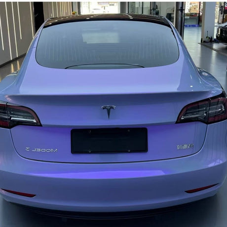 A rear view of a Tesla Model 3 with Gray to Purple Charm Glossy SelfRepair Nano-Coated PPF, tinted windows, no license plate, parked indoors in a bright showroom.