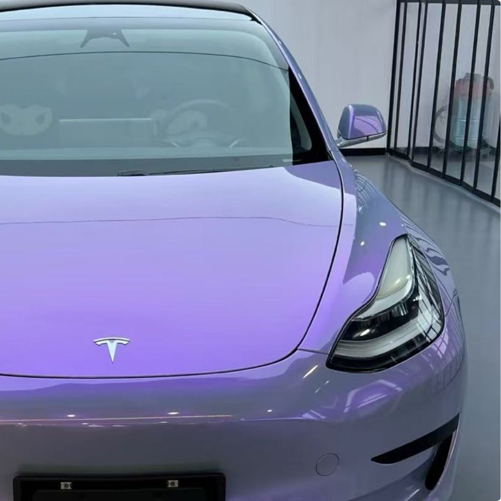 A front view of a shiny Tesla in Gray to Purple Charm Glossy SelfRepair Nano-Coated PPF shows vibrant color effects and protective film, with reflections on the hood and windshield, parked indoors by a black metal railing.