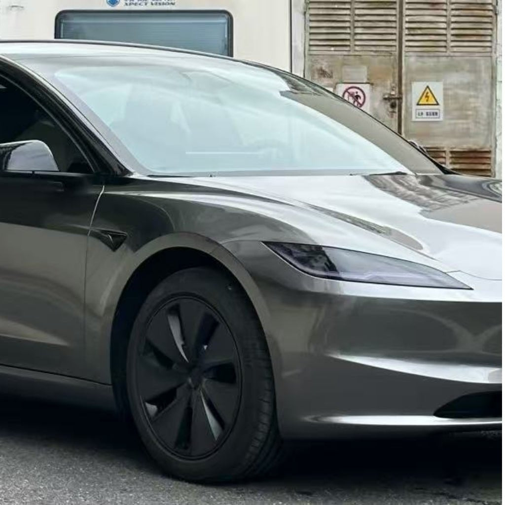 A sleek, silver electric car with black wheels and minimalist headlights is parked on the street, protected by Gray Liquid Metallic Glossy SelfRepair Nano-Coated PPF. In the background, a building displays warning signs and closed shutters.