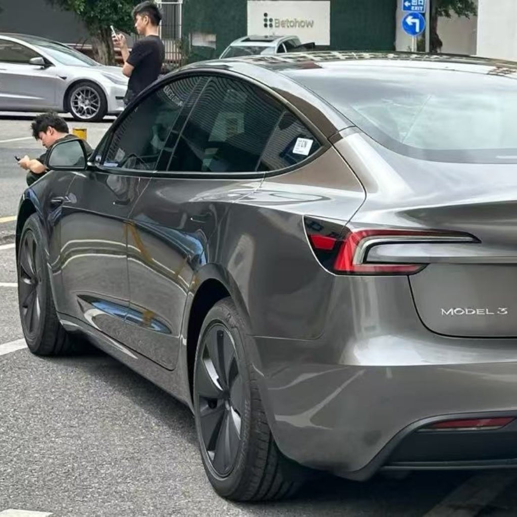 A metallic gray Tesla Model 3 protected by Gray Liquid Metallic Glossy SelfRepair Nano-Coated PPF is parked on the street. One person crouches near the front while another stands nearby, possibly taking a photo with a phone.