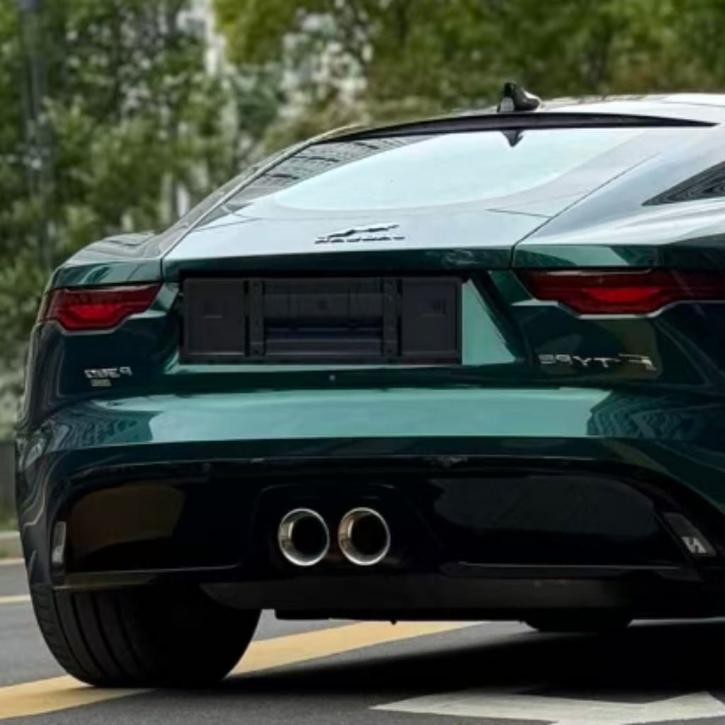 A close-up of the rear end of a dark green Jaguar F-Type in Diamond British Green Glossy SelfRepair Nano-Coated PPF, highlighting dual exhaust pipes, taillights, and a blank license plate area with trees in the background.