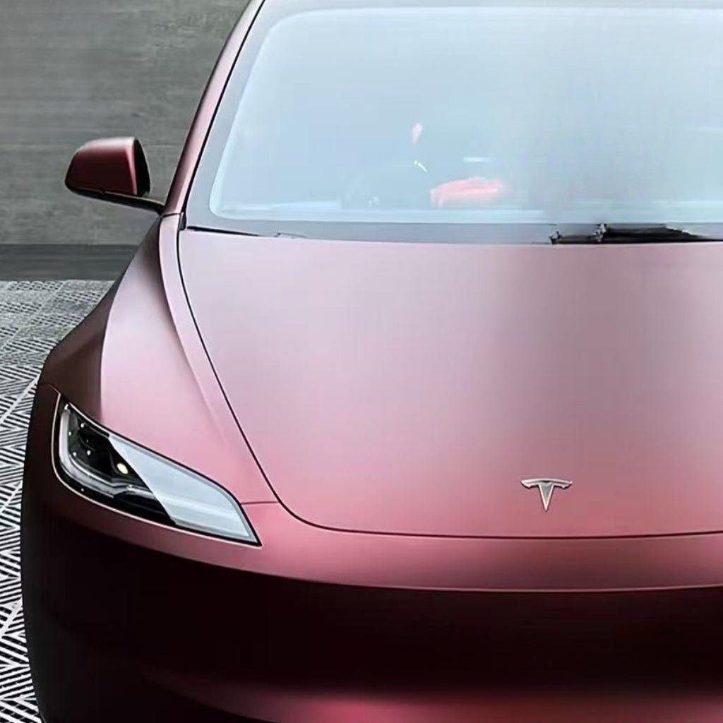 A close-up of a Tesla's hood, left headlight, and logo showcases the Dragon Blood Red Matte SelfRepair Nano-Coated PPF, parked on a patterned surface with a concrete wall in the background.