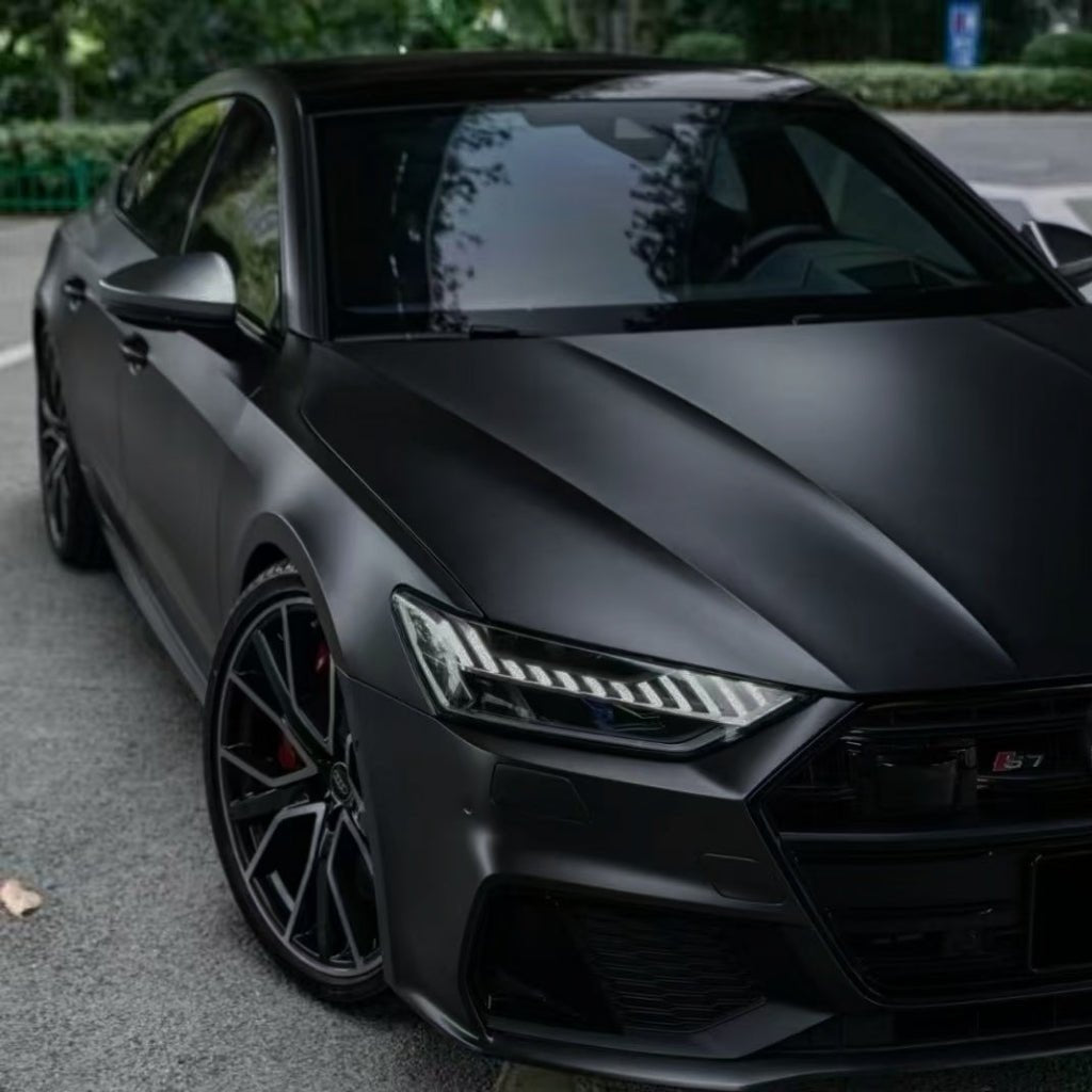 A sleek black sports car with a matte finish is parked by the roadside, surrounded by greenery. It features modern headlights, stylish black wheels, and is protected by Phantom Black Matte SelfRepair Nano-Coated PPF for lasting elegance.