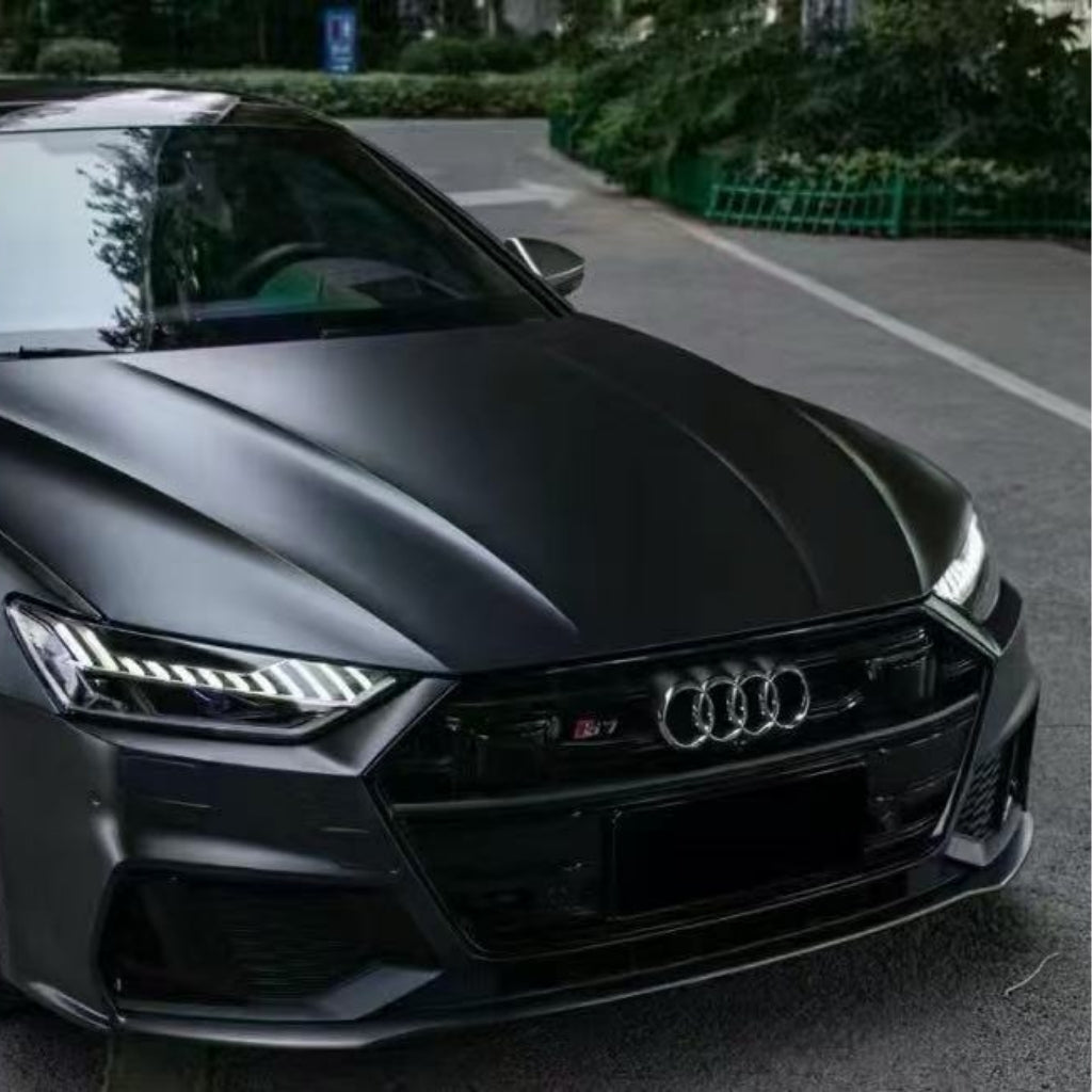 A close-up of the front of a black Audi RS7 parked on a street, featuring its sleek hood protected by Phantom Black Matte SelfRepair Nano-Coated PPF, LED headlights, and the distinctive Audi logo on the grille.