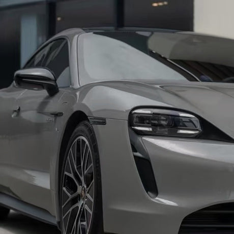 Close-up shot of the front left side of a sleek silver sports car with modern LED headlights and alloy wheels, featuring Porsche Gray Matte SelfRepair Nano-Coated PPF, parked in front of a contemporary building.