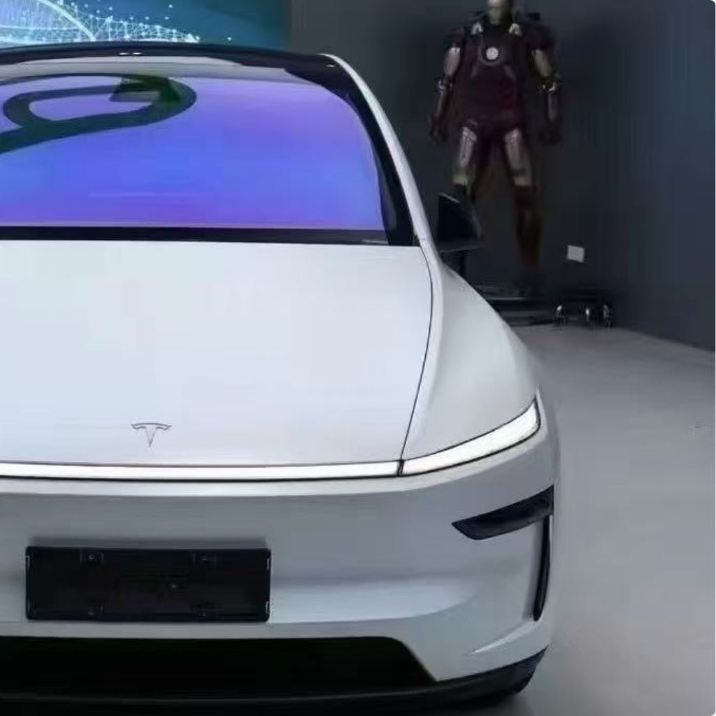 A white Tesla with a sleek, modern design displays indoors, its windshield reflecting a digital screen. It features Transparent Sparkle Matte SelfRepair Nano-Coated PPF for top paint protection. An Iron Man suit stands nearby in the background.