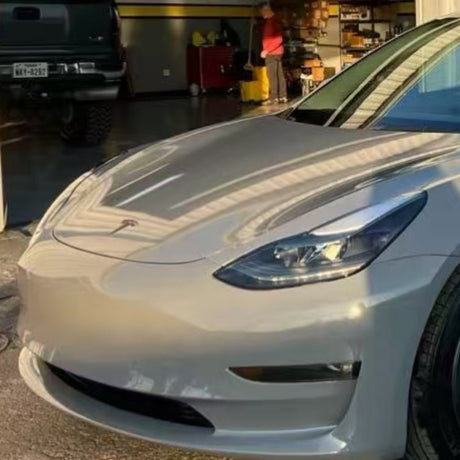 A silver Tesla Model 3 with Nardo Gray Matte SelfRepair Nano-Coated PPF is parked outside a garage, where part of a black pickup truck is visible in the background and a person stands near shelves inside.