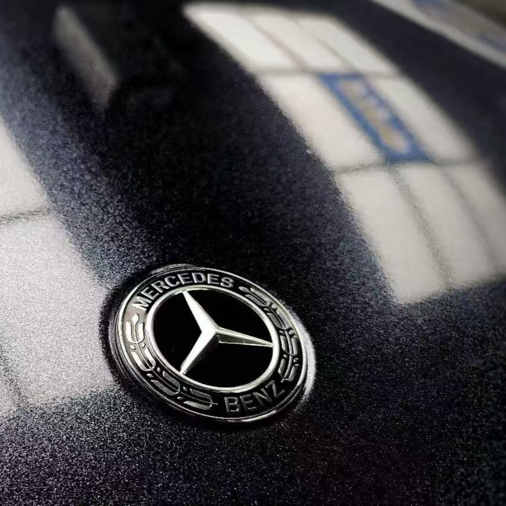 Close-up of a Mercedes-Benz logo on a hood wrapped in Midnight Black Sparkle Glossy SelfRepair Nano-Coated PPF, as bright window reflections form geometric patterns on the dark, shiny surface.