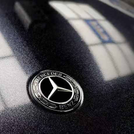 Close-up of a Mercedes-Benz logo on a hood wrapped in Midnight Black Sparkle Glossy SelfRepair Nano-Coated PPF, as bright window reflections form geometric patterns on the dark, shiny surface.