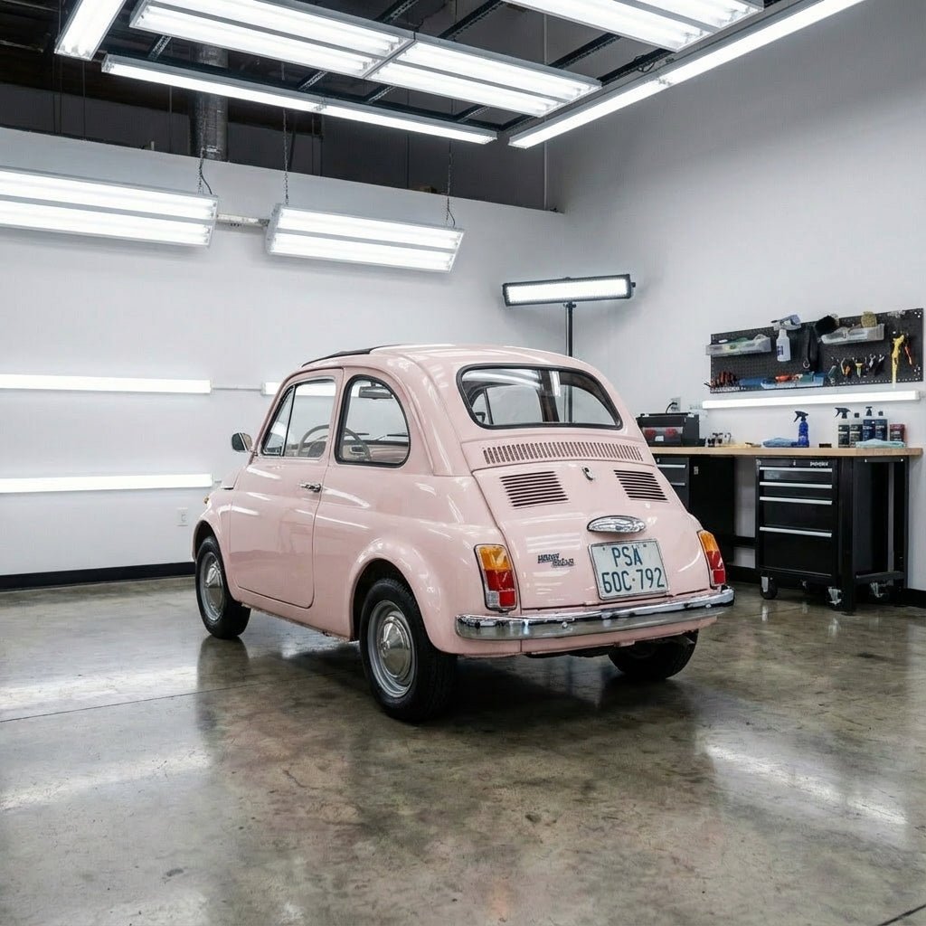 A vintage pink Fiat 500, wrapped in Pearl Blossom Pink Glossy SelfRepair Nano-Coated Nexthor™ PPF, is parked inside a modern garage with white walls, polished concrete floor, and side workbenches filled with tools.