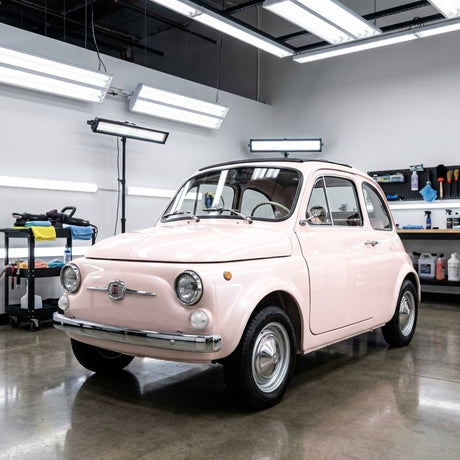 A vintage light pink Fiat 500, protected by Pearl Blossom Pink Glossy SelfRepair Nano-Coated Nexthor™ PPF, is parked in a modern garage surrounded by bright lights and detailing equipment.