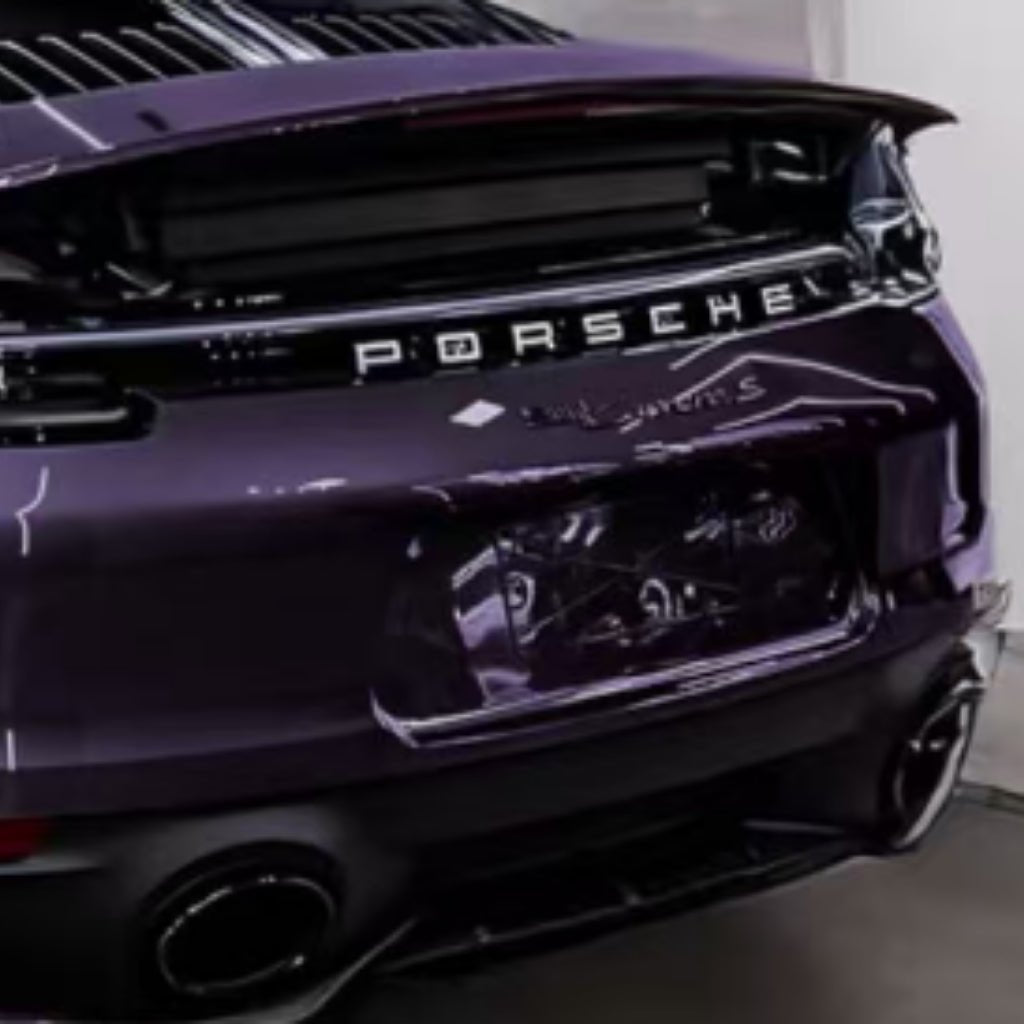 A close-up of the rear of a purple Porsche Taycan Turbo S parked indoors, showing the Porsche logo, raised spoiler, and taillights protected by Diamond Provence Purple Glossy SelfRepair Nano-Coated PPF.