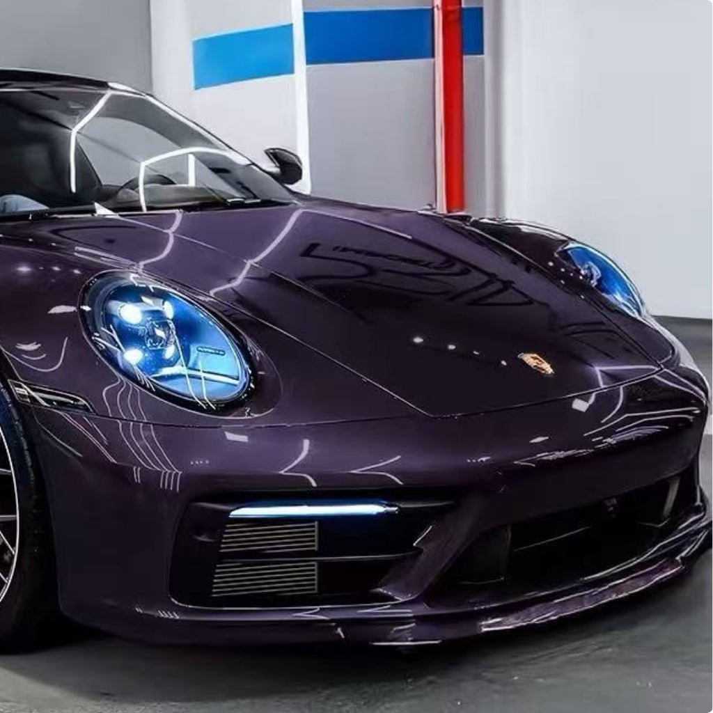 A close-up of a dark purple Porsche with blue-tinted headlights displays Diamond Provence Purple Glossy SelfRepair Nano-Coated PPF, offering ochrana karosérie while parked indoors by a white wall with a blue stripe and red pipe.